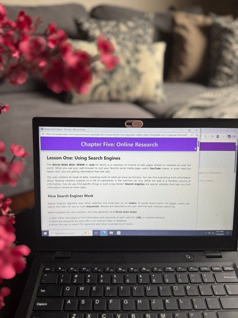 A laptop displaying a lesson on Online Research from CompuScholar's Tech Essentials curriculum, featuring a purple header and educational content about using search engines, with decorative flowers in the foreground.