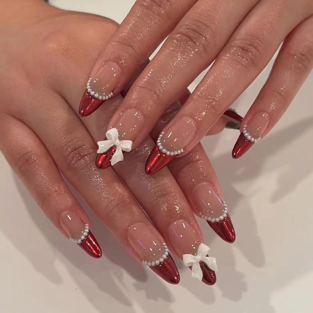 Close-up of two hands showcasing long, pointed nails decorated with red tips, white bows, and small pearl accents.