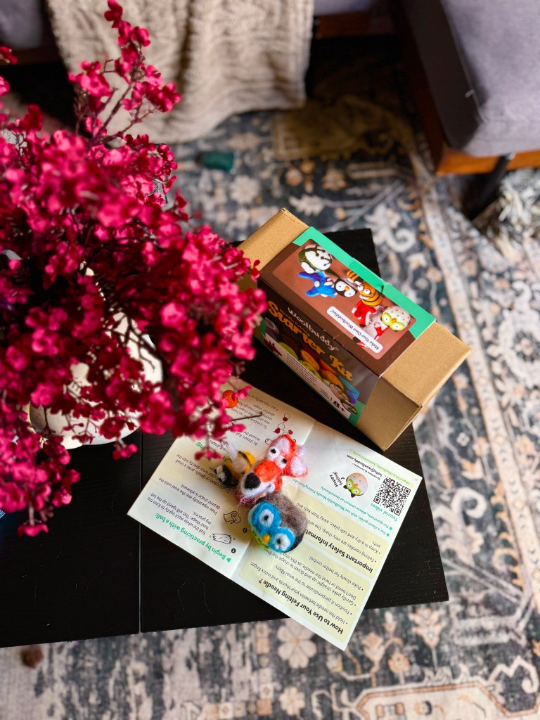 A vibrant Woolbuddy Needle Felting Starter Kit displayed on a table, alongside colorful felted animal figures and a flower arrangement. The kit box is partially open, revealing the contents and instructional materials.
