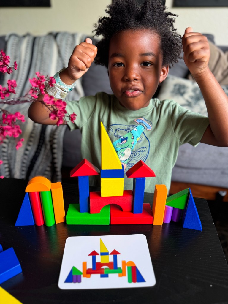 A young child with curly hair smiles and raises their fists in excitement, sitting at a table with colorful building blocks arranged in a creative structure. A card showing a similar building design is placed in front of them.