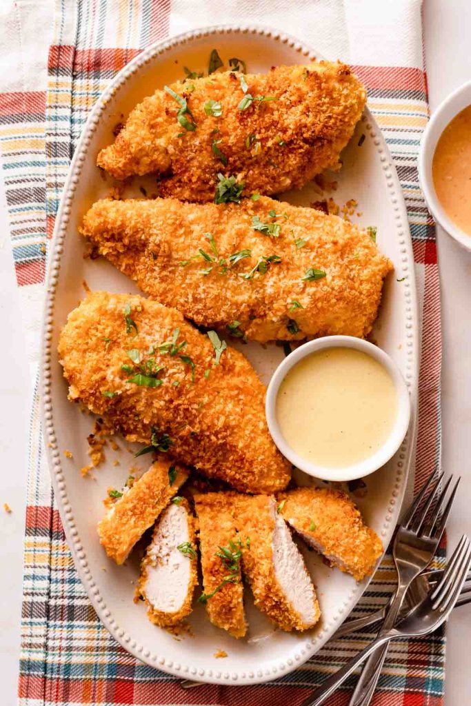 A platter of crispy panko baked chicken breasts garnished with fresh herbs, accompanied by a small bowl of dipping sauce.
