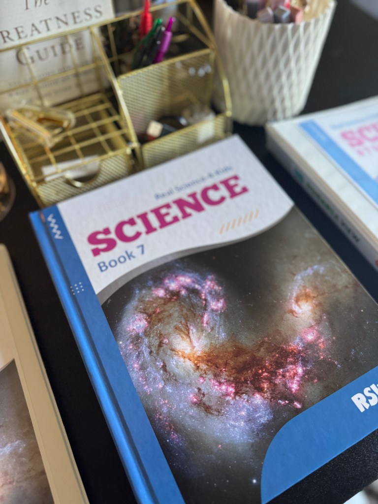 A close-up of the 'Real Science 4 Kids Book 7' textbook featuring a colorful image of a galaxy on its cover, placed on a desk with pens and stationery accessories.