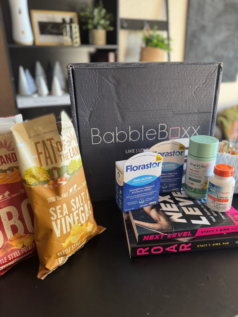 A flat lay image showcasing a BabbleBoxx filled with wellness products, including bags of 'Fat of the Land' chips, a box of 'Florastor' probiotics, and two books, 'ROAR' and 'Next Level' by Dr. Stacy Sims, placed on a dark surface.