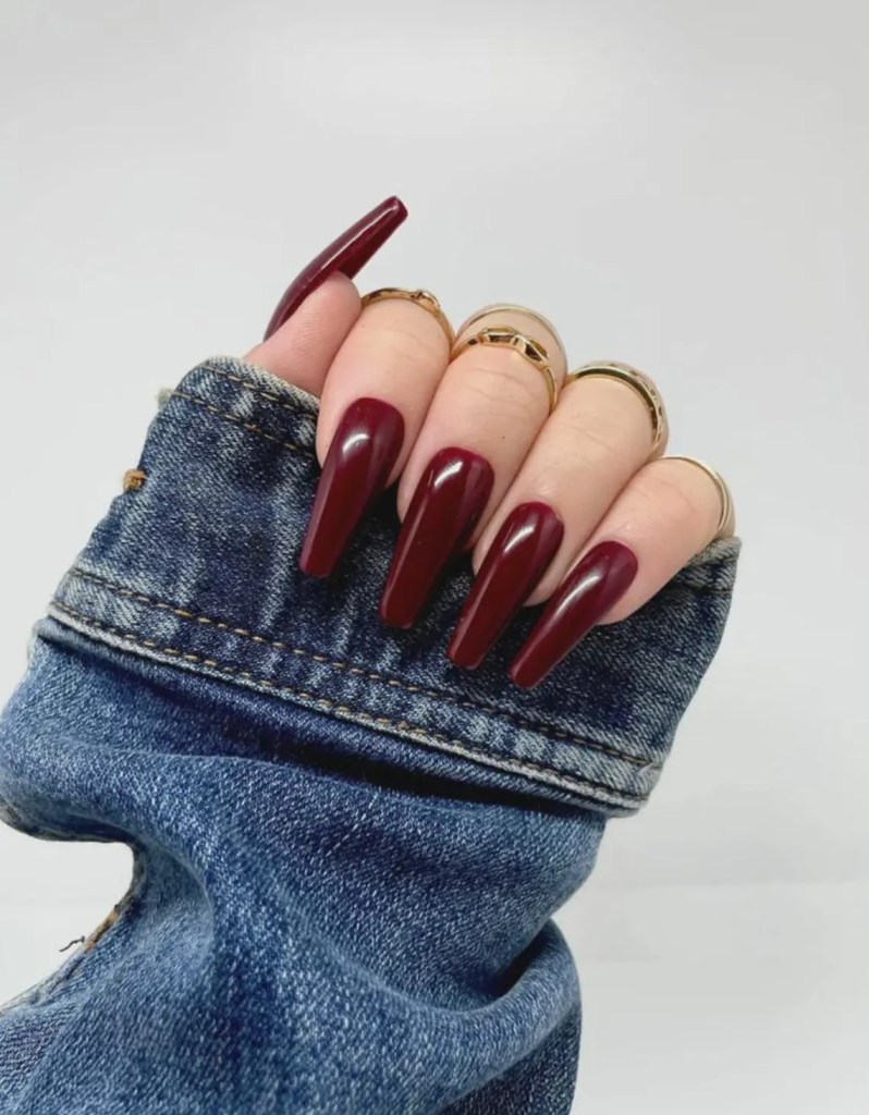 A close-up of a hand with long, pointed nails painted in a dark burgundy color, adorned with several gold rings, resting on a denim jacket sleeve.