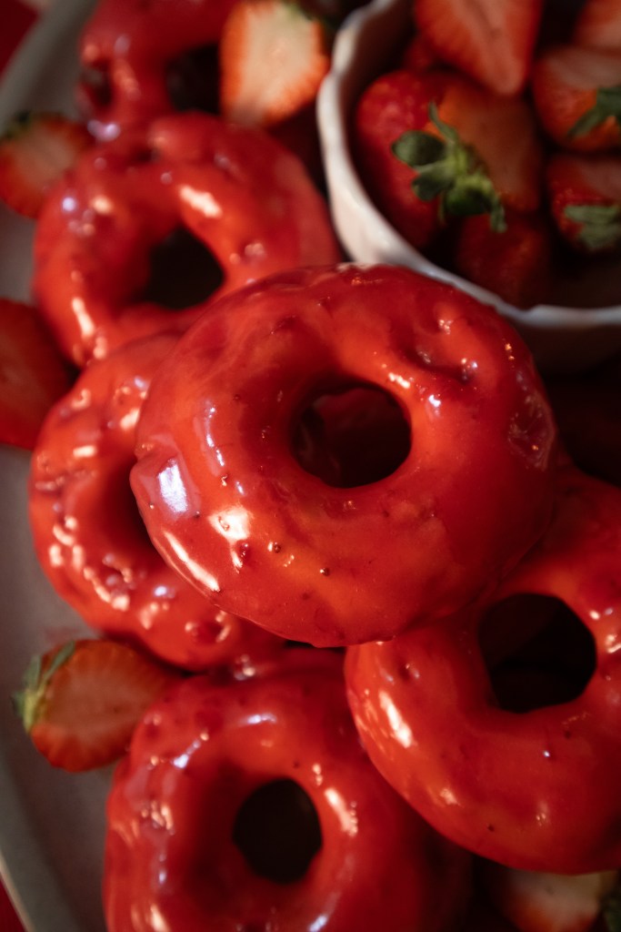 A close-up of glazed red vegan strawberry donuts surrounded by fresh strawberries on a plate.
