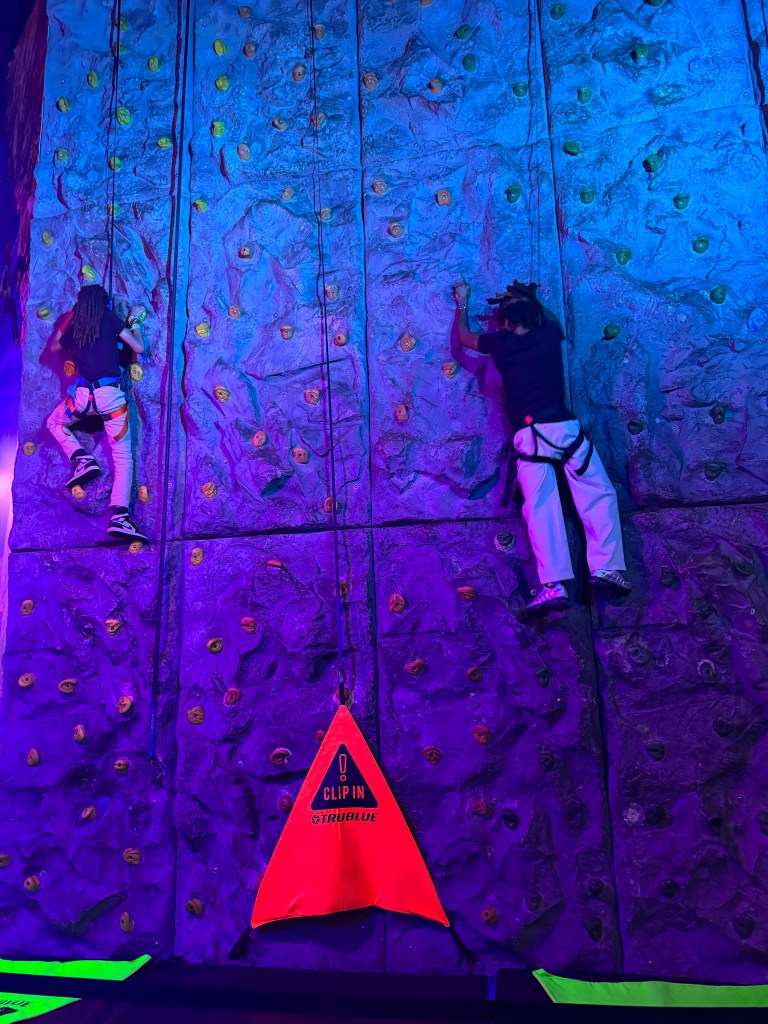 Two climbers ascending a brightly colored indoor rock climbing wall with safety harnesses, under purple and blue lighting.
