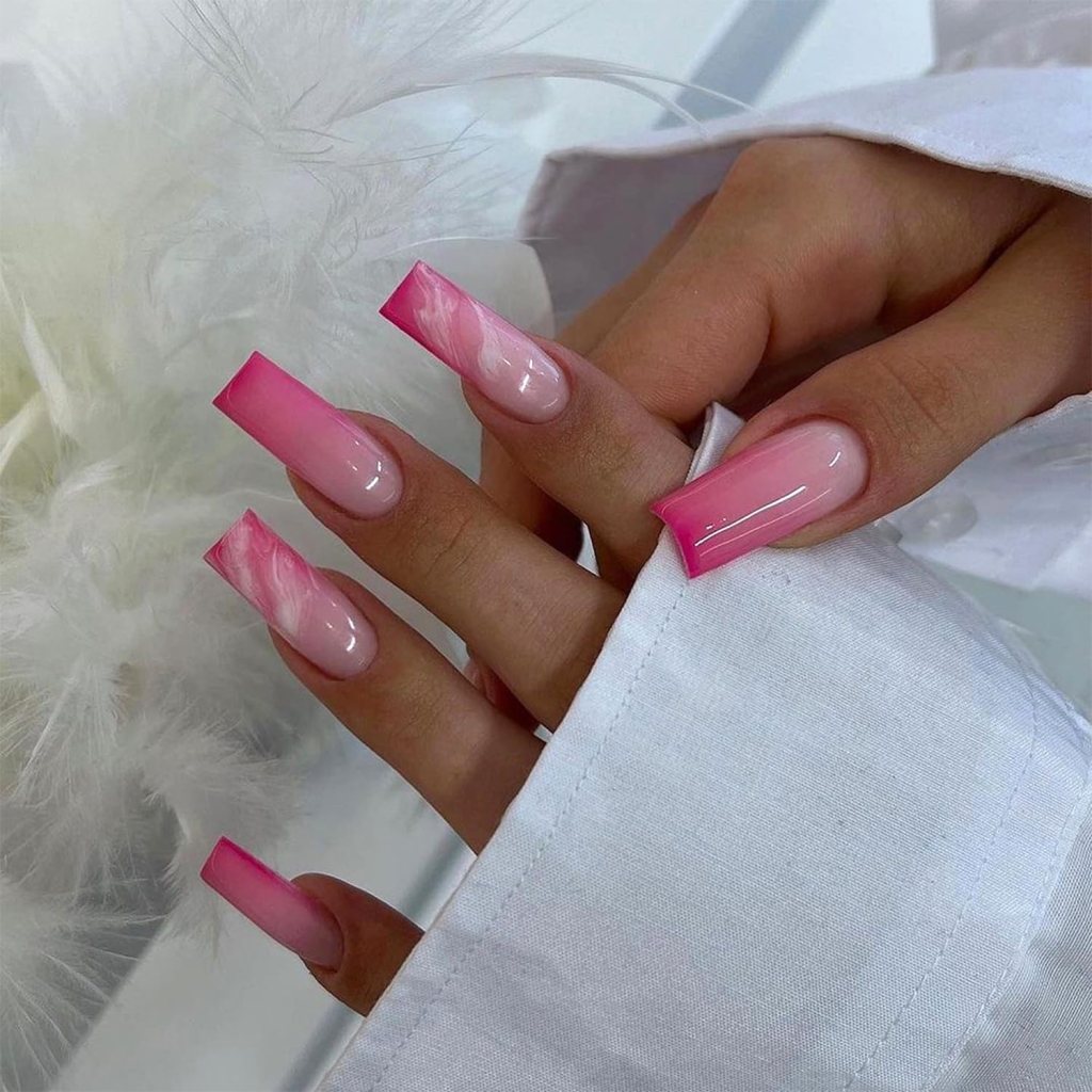 A close-up of manicured nails featuring a pink ombre design with square tips, blending from light pink to deep pink, resting on a white fabric.