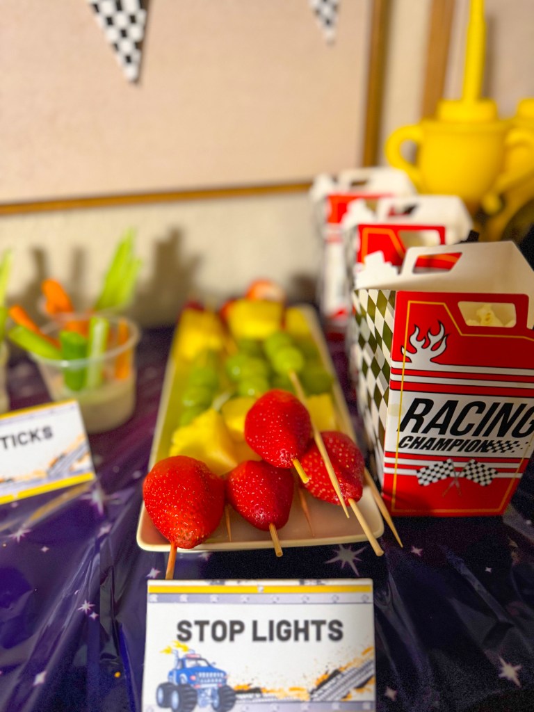 Monster truck birthday party food idea with stop light fruit skewers made with strawberries, pineapple, and grapes on a racing themed snack table.