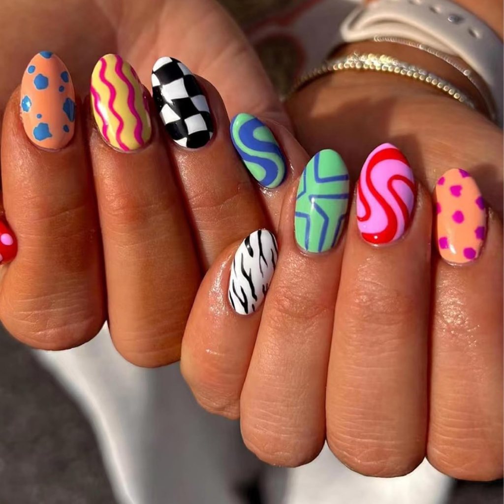 A close-up of a hand displaying colorful, patterned nails featuring various designs like polka dots, wavy lines, and checkerboard.