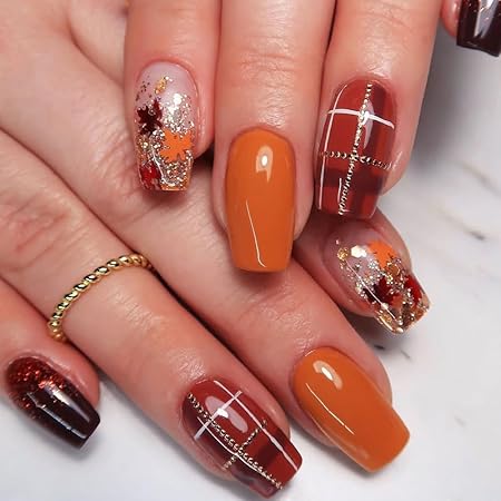 A hand showing autumn-themed nails featuring burnt orange, brown, and gold accents, with a mix of metallic and plaid designs.