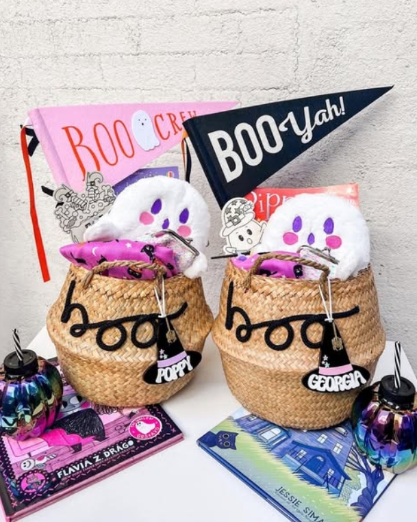 Two personalized woven baskets labeled 'Poppy' and 'Georgia' filled with Halloween plush toys and pink accessories, accompanied by 'Boo Crew' and 'Boo Yah!' pennants and colorful books.