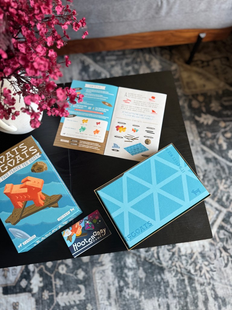 A tabletop game 'That Floats Yer Goats' displayed on a dark wooden table alongside colorful instruction brochures and a business card for Hootenanny Games, with a decorative pink flower arrangement in the background.