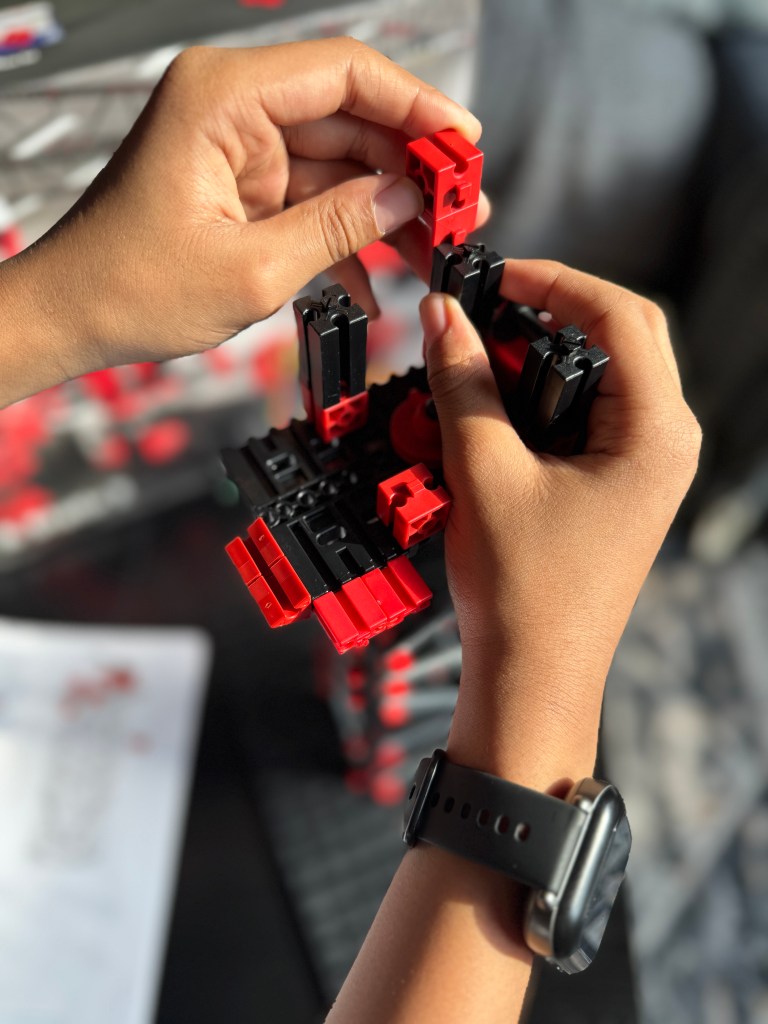 Two hands assembling a building set with red and black pieces, focusing on the intricate connections and construction process.