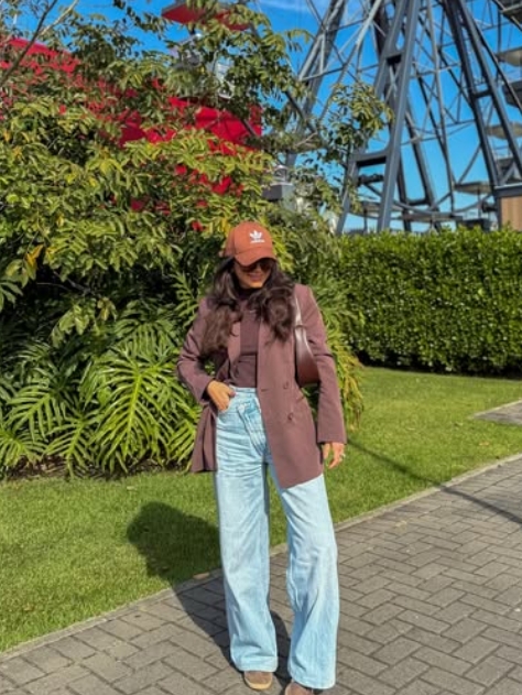 A woman wearing a maroon blazer, a brown cap, and light blue wide-leg jeans stands outdoors, surrounded by greenery and modern architecture.