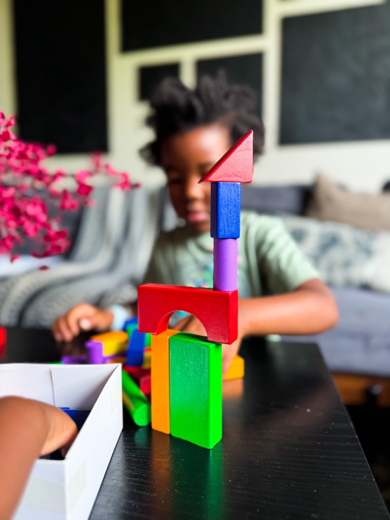A young child is building a colorful tower with wooden blocks on a table, with various block shapes and colors in the foreground and a blurred background of a cozy living room.