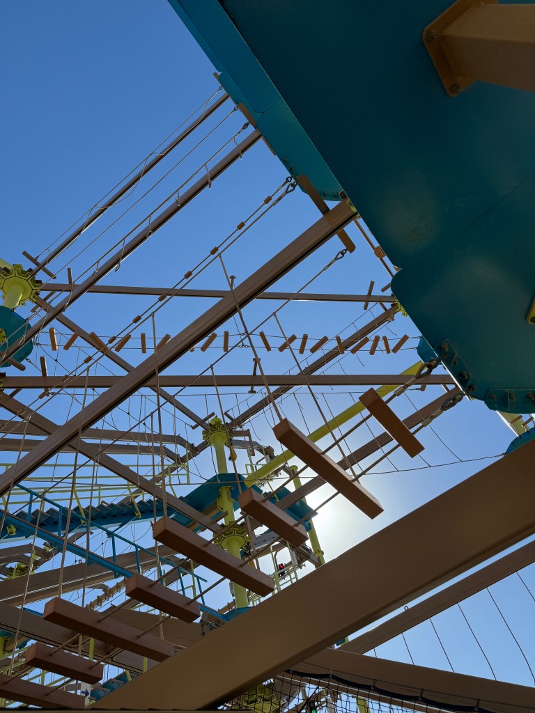View from underneath a high ropes course, showcasing a network of beams and platforms against a clear blue sky.
