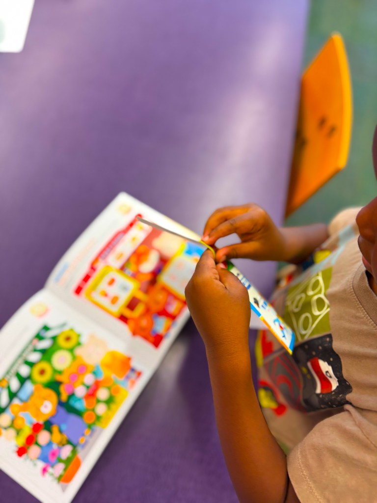 A child holding a colorful book with stickers, sitting at a table covered in purple material.