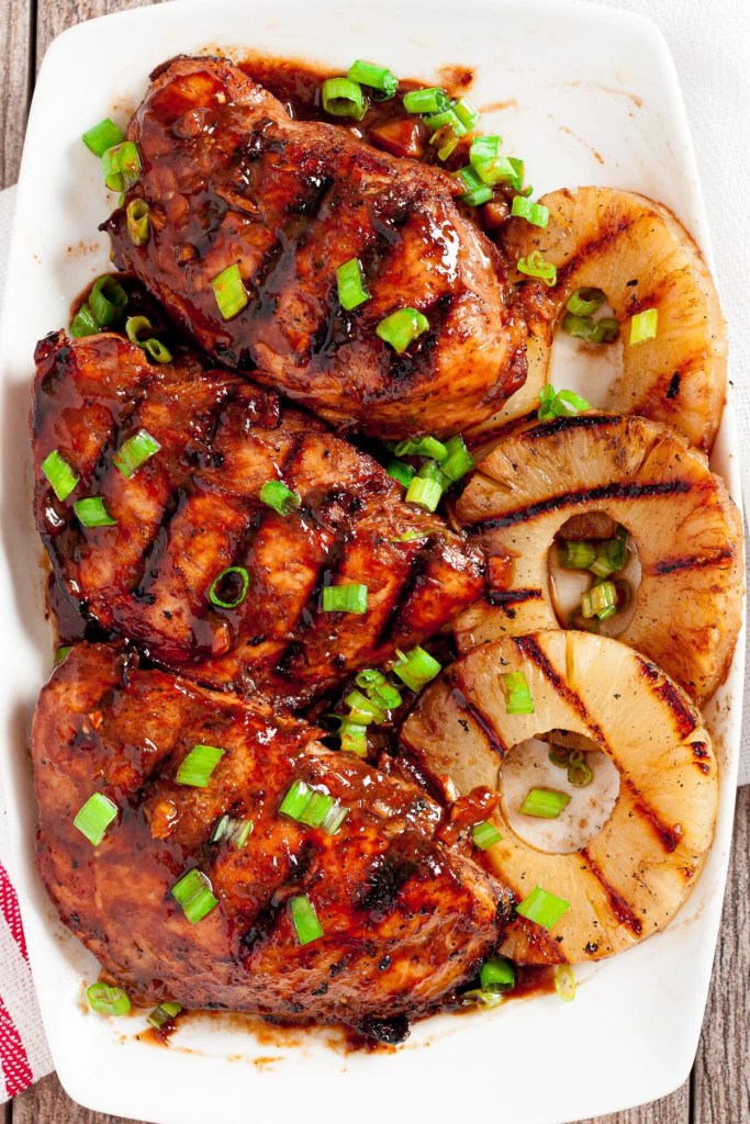 Grilled chicken breasts coated in a dark sauce, garnished with chopped green onions, served with grilled pineapple rings on a white plate.