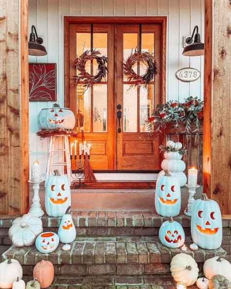 A cozy porch decorated for fall, featuring wooden double doors, playful blue and white pumpkins with jack-o'-lantern faces, and lush greenery. Two wreaths adorn the doors, creating a warm and inviting autumn entrance.
