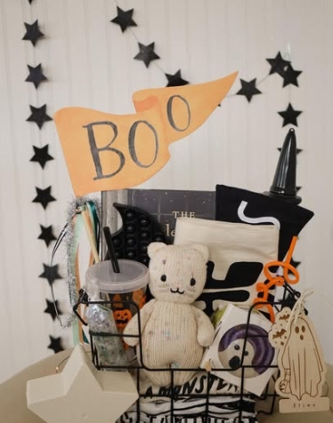 A Halloween-themed basket filled with a stuffed toy, snacks, craft supplies, and decorations, featuring a flag that says 'BOO' and surrounded by star decorations.