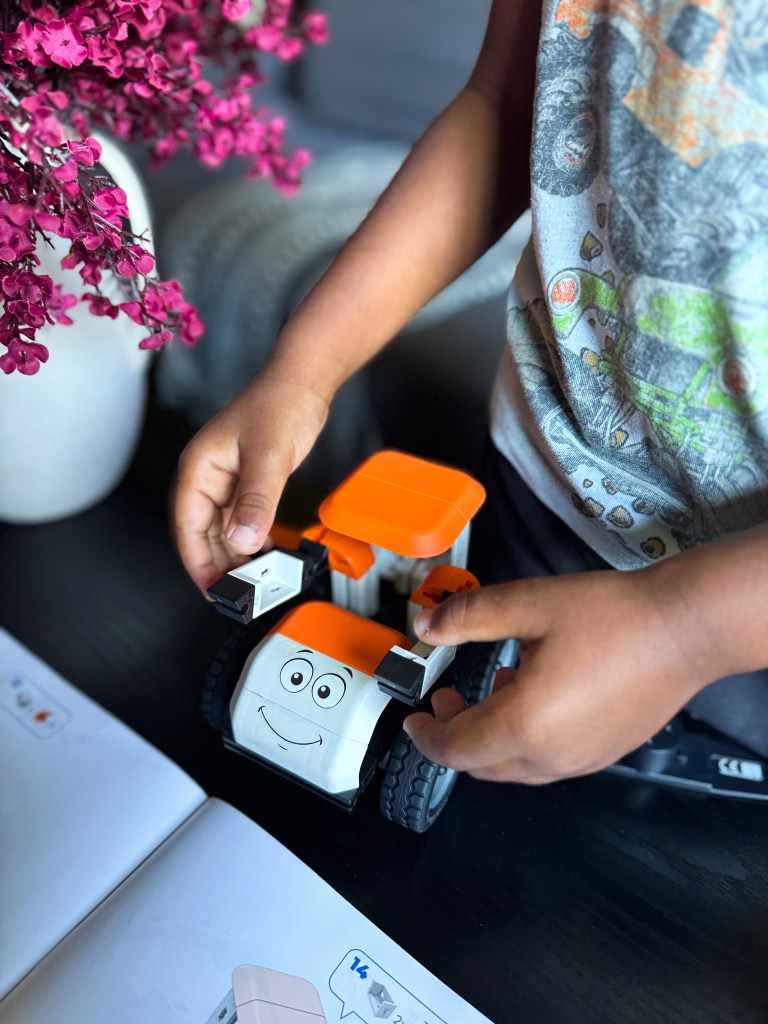 A child assembling a toy vehicle with a smiling face, holding the vehicle's parts. In the background, pink flowers and an instruction manual are visible.