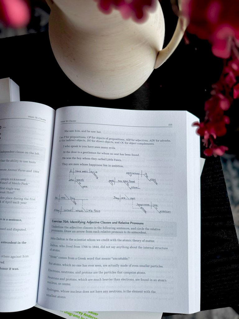 A close-up view of an open textbook page featuring notes on adjective clauses and relative pronouns, with a decorative vase and flowers in the background.