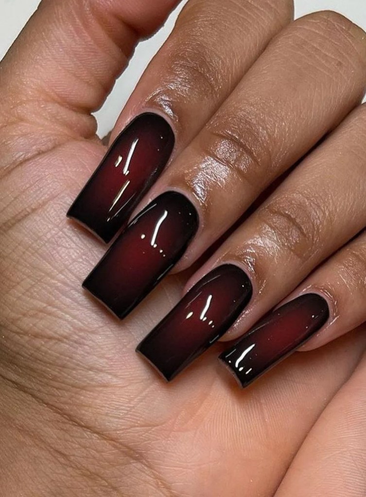 A close-up image of a hand displaying long, glossy nails painted in a gradient color that transitions from deep red at the tips to black at the base.