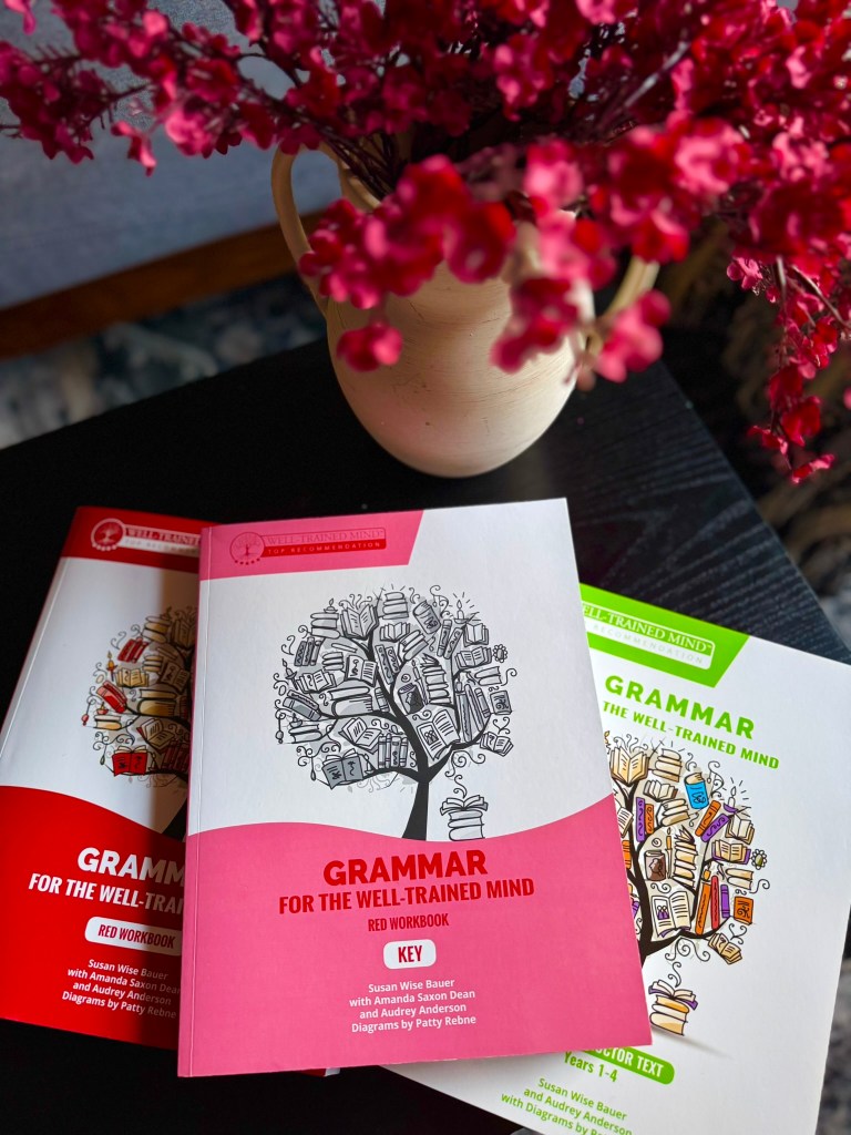 A close-up of a vase with pink flowers beside three grammar workbooks titled 'Grammar for the Well-Trained Mind', featuring a tree design filled with books. The workbooks include a red workbook and a key edition.