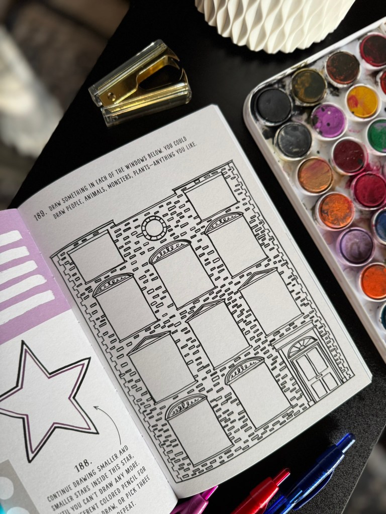 Open art book page featuring a prompt to draw in windows, surrounded by artistic supplies like watercolors, a stapler, and pens.