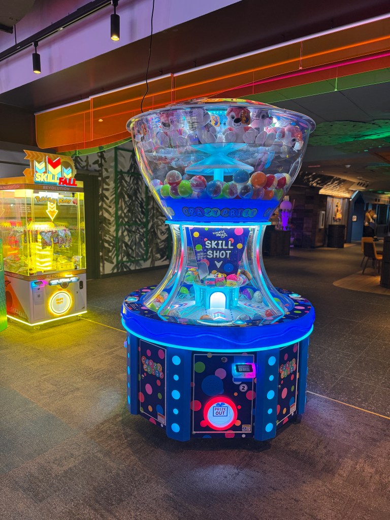 A colorful arcade game machine featuring a candy and plush toy dispenser, located in the lively Northern Lights Arcade at Great Wolf Lodge in Grapevine, TX.