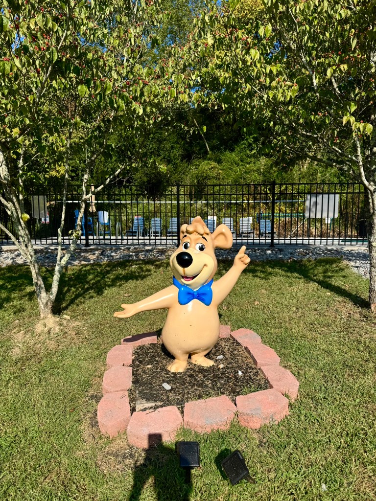 A cheerful cartoon bear statue wearing a blue bow tie, standing in a bed of mulch surrounded by low decorative bricks, with green trees and a fence in the background.