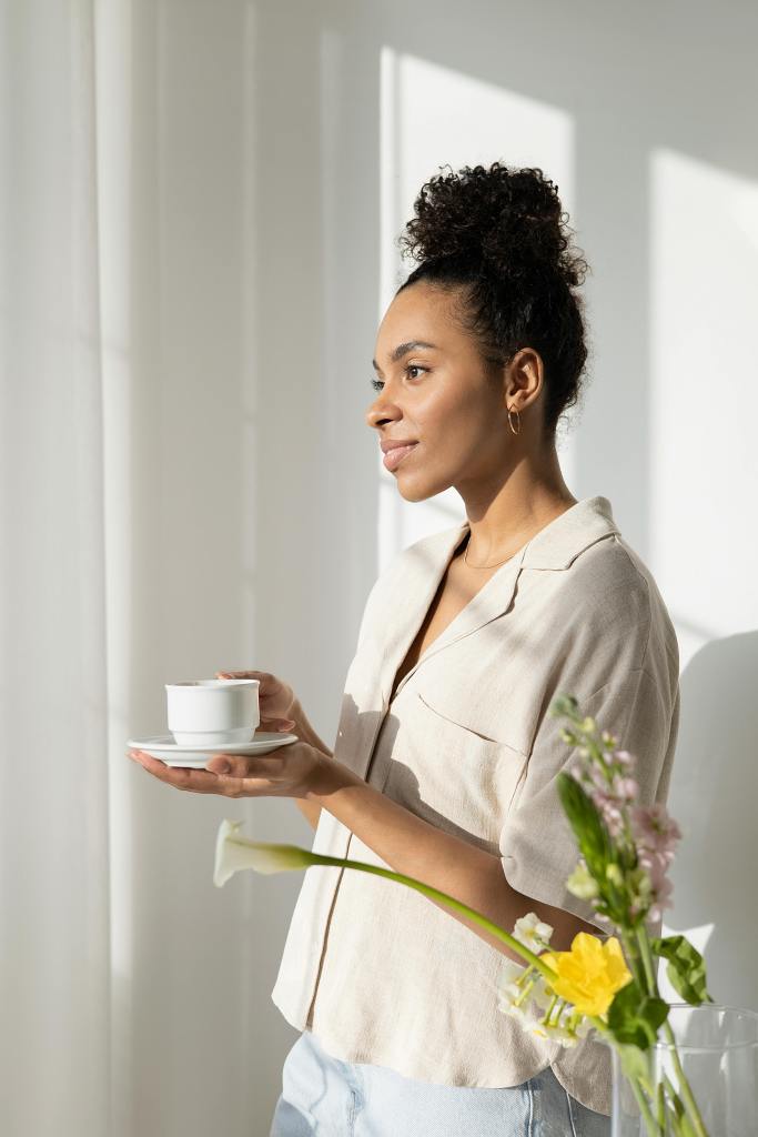 A woman standing by a window holds a small cup and saucer while looking thoughtfully outside, surrounded by flowers and soft natural light.