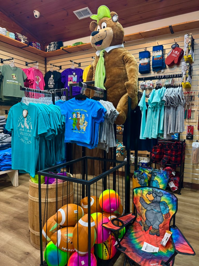 A retail store display featuring a large plush bear wearing a green hat and tie, surrounded by various colorful t-shirts and children's toys including balls and chairs.