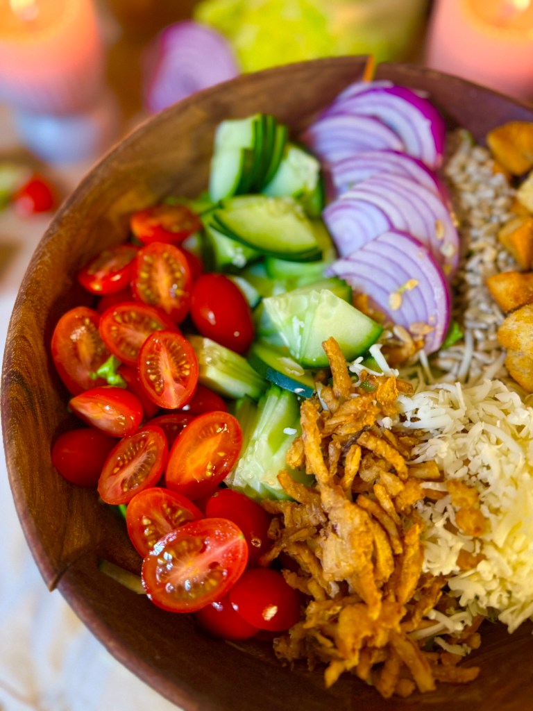 A wooden bowl filled with a colorful salad featuring cherry tomatoes, sliced cucumbers, red onions, fried onions, and white rice.