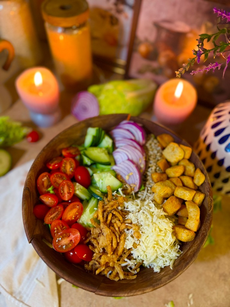 A wooden bowl filled with a colorful salad featuring cherry tomatoes, cucumber slices, red onion rings, fried onions, grated cheese, and small crunchy snacks, with candles and decorative elements in the background.