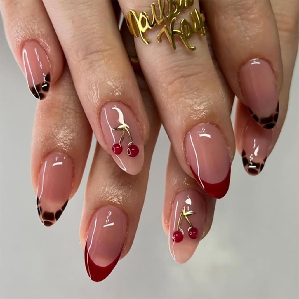 A close-up of manicured hands showcasing a stylish nail art design with a mix of nude, red, and leopard print patterns. Some nails feature small cherry embellishments and a gold ring.