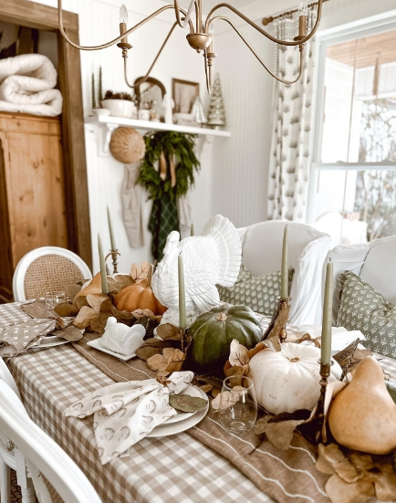 A cozy fall tablescape featuring a mix of pumpkins in various colors, greenery, and soft candlelight, arranged on a checkered tablecloth with elegant tableware.