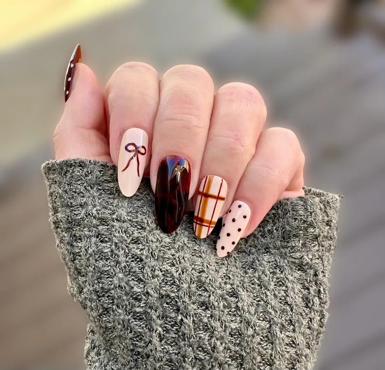 A hand displaying festive Thanksgiving nail art featuring a mix of burnt orange, chocolate brown, and neutral tones with designs like a bow, plaid, and polka dots, set against a cozy knitted sweater.