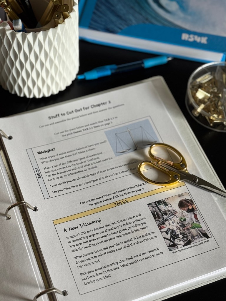 A close-up view of an open science curriculum book showing cut-out instructions for a chapter, with a pair of golden scissors and stationery in a decorative holder next to it, and a blue pen on the side.