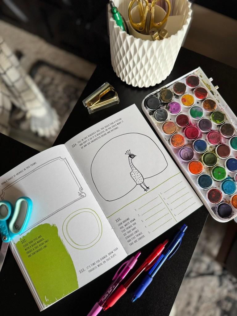 A creative workspace featuring an open art prompt book from 'My Year of Art,' surrounded by colored pens, scissors, a stapler, and a watercolor paint palette.