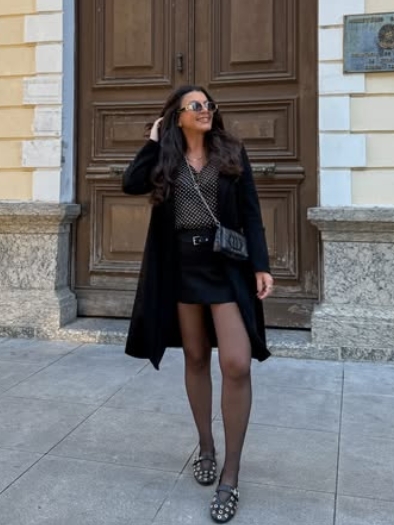 A woman wearing a black coat over a patterned top and mini skirt, paired with black tights and stylish sandals, poses in front of a wooden door.