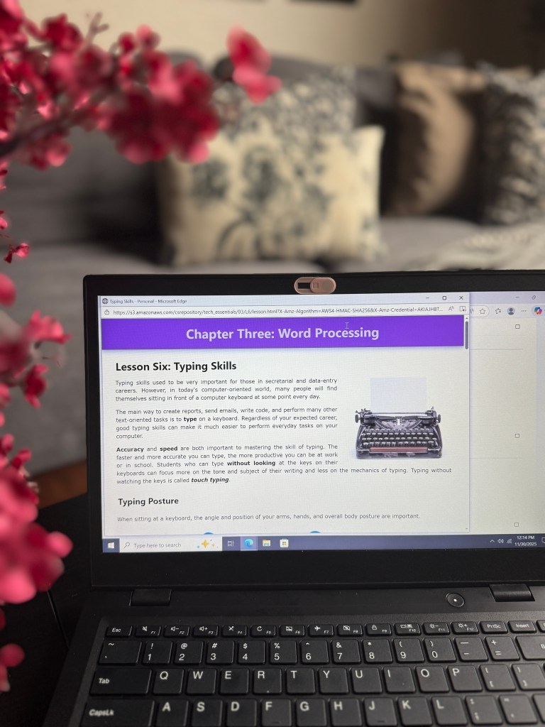 A laptop displaying an online lesson titled 'Chapter Three: Word Processing' with a focus on typing skills, alongside a blurred pink flower arrangement in the foreground.