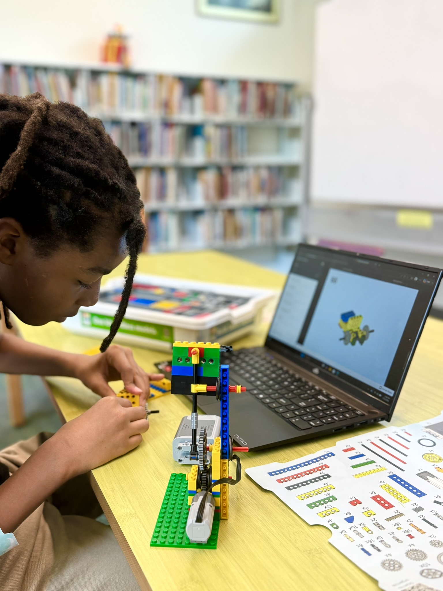 Child working on a LEGO robotics project with instructions and a laptop in a library