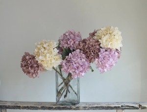 A vase filled with hydrangeas in shades of pink, lavender, and cream, sitting on a rustic wooden surface with a neutral background.