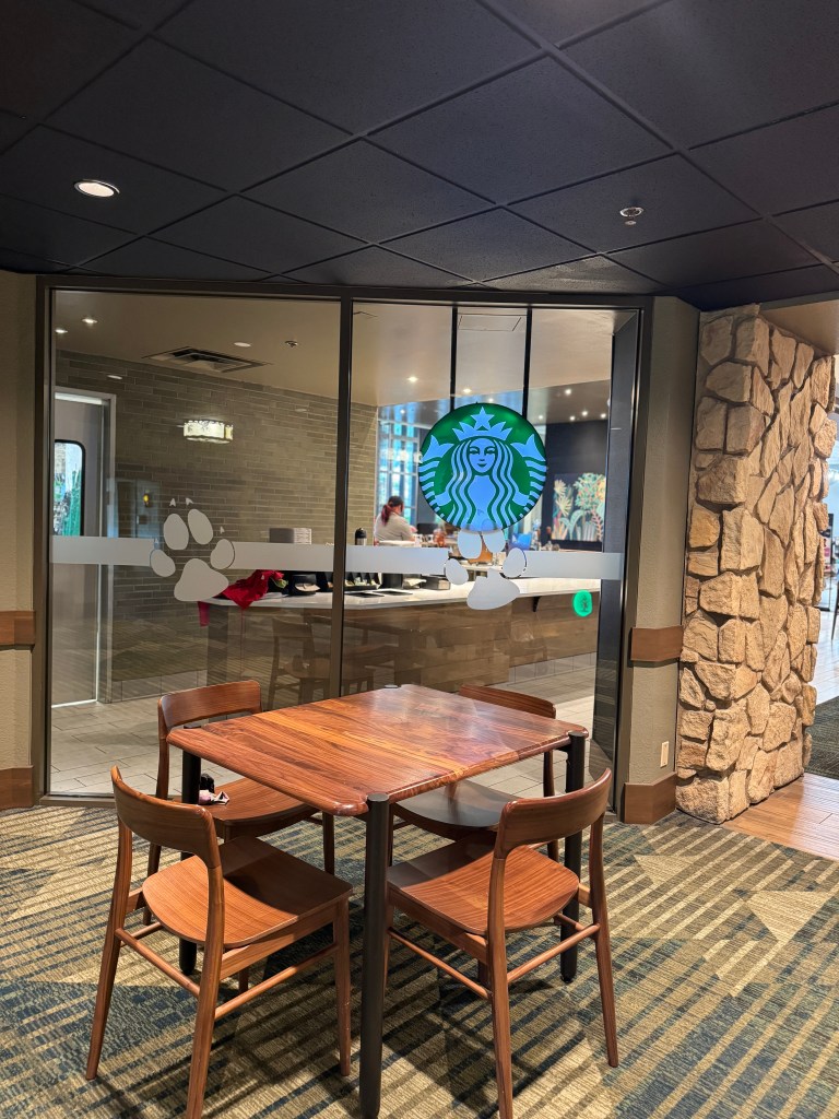 Entrance of a Starbucks located inside Great Wolf Lodge, featuring a prominent logo and paw print decals on the glass door, with a wooden table and chairs in the foreground.