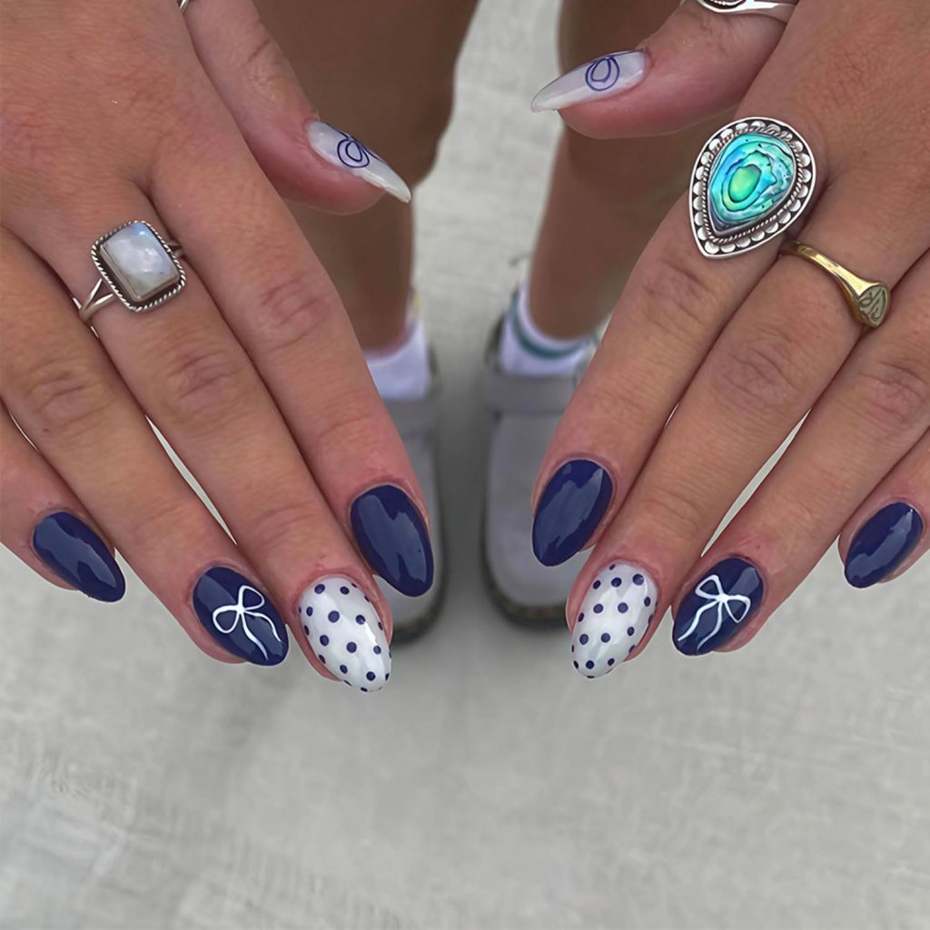 Navy blue and white almond nails with polka dots, bow accents, and glossy coquette-inspired nail art.