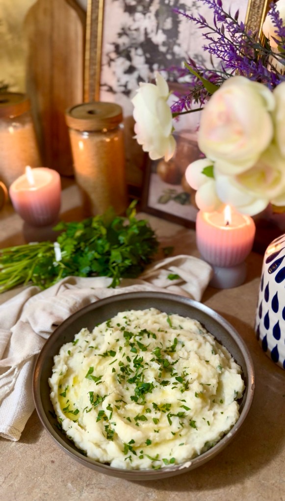 A bowl of creamy mashed potatoes topped with fresh herbs, surrounded by candles, decorative jars, and flowers.