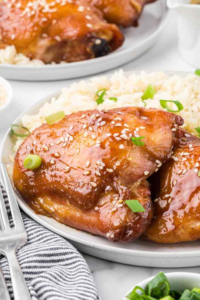 A close-up of glazed chicken thighs served over a bed of rice, garnished with green onions and sesame seeds, on a white plate with a striped napkin and a small bowl of green onions in the background.