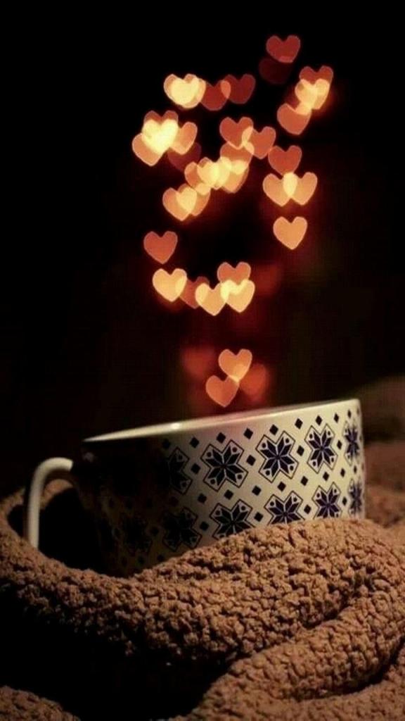A cozy mug with a decorative pattern, wrapped in a warm blanket, surrounded by twinkling heart-shaped bokeh lights against a dark background.