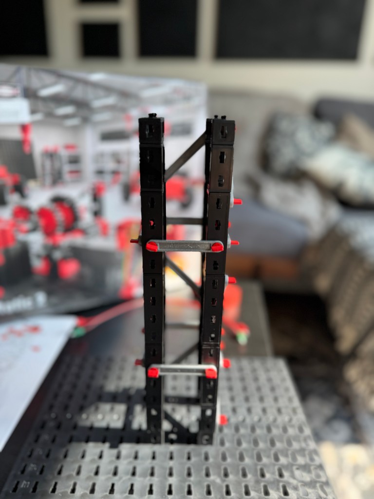 A completed structure made from a Fischer Technik STEM kit, showcasing intricate black and red components on a textured surface, with a blurred background of a workshop or play area.